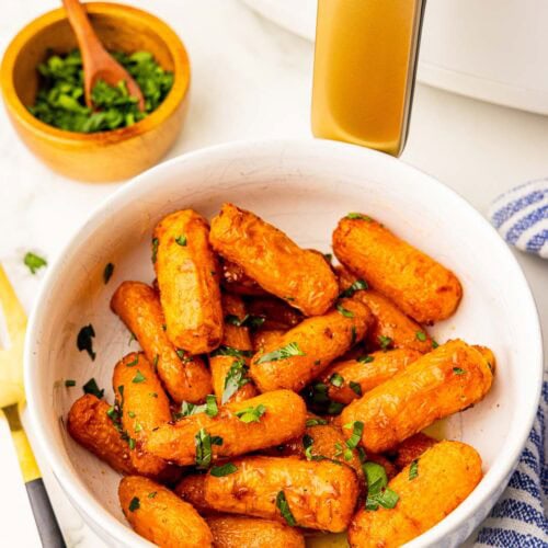 overhead view of white bowl of air fried baby carrots sprinkled with parsley, with bowl of parsley and white air fryer in background