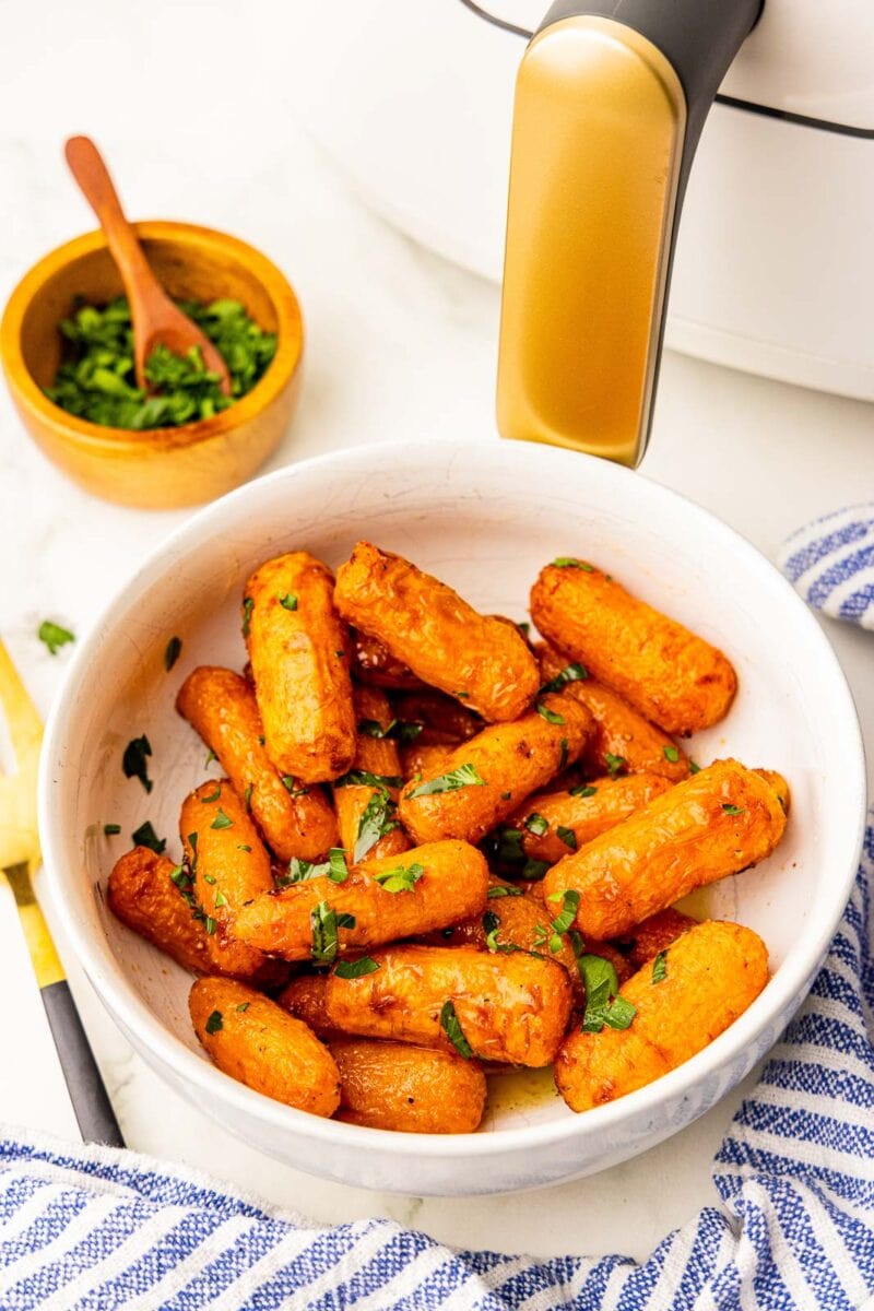 overhead view of white bowl of air fried baby carrots sprinkled with parsley, with bowl of parsley and white air fryer in background