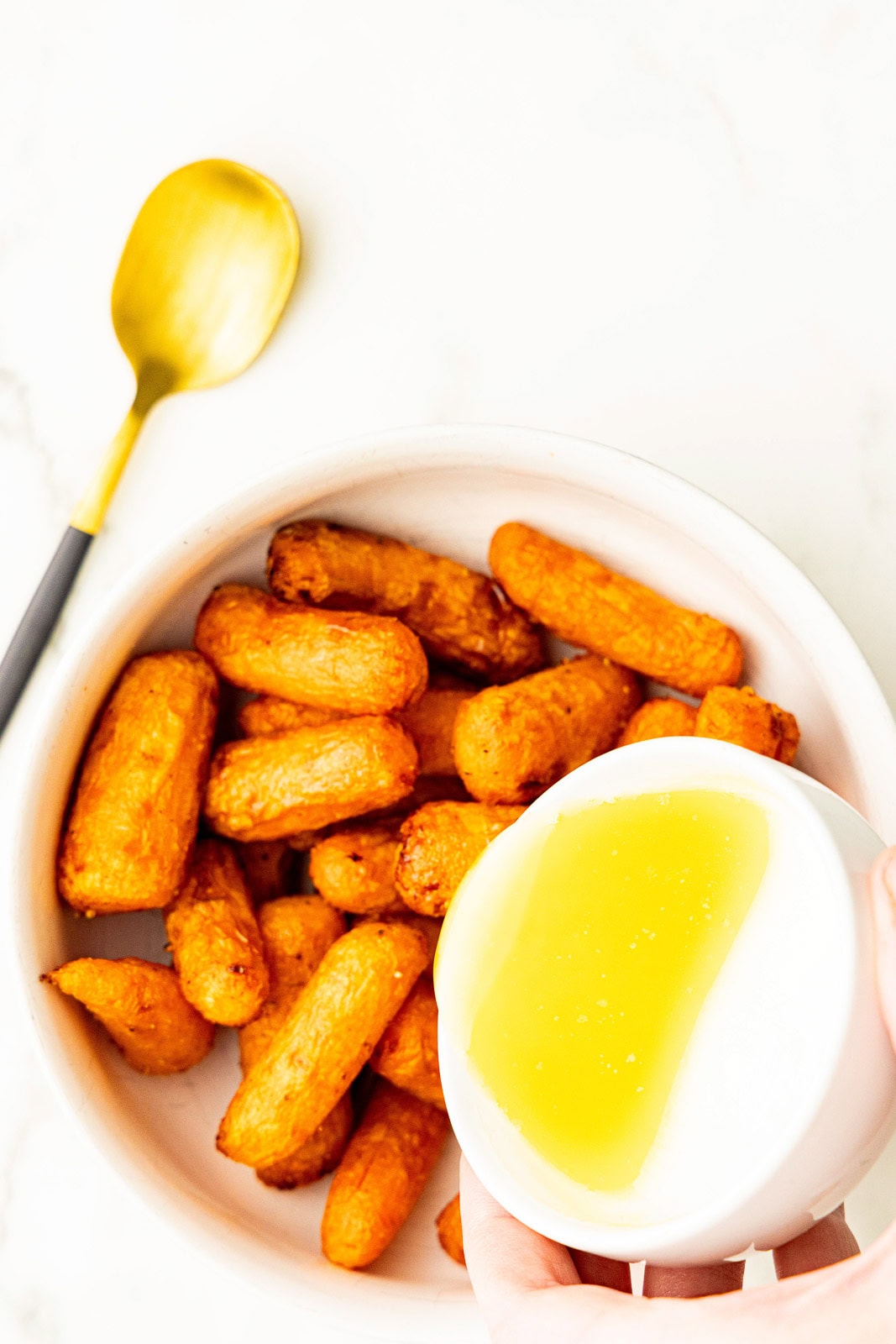 overhead view of large white bowl of air fried baby carrots with small bowl of honey/butter mixture being poured over carrots, with gold spoon on side