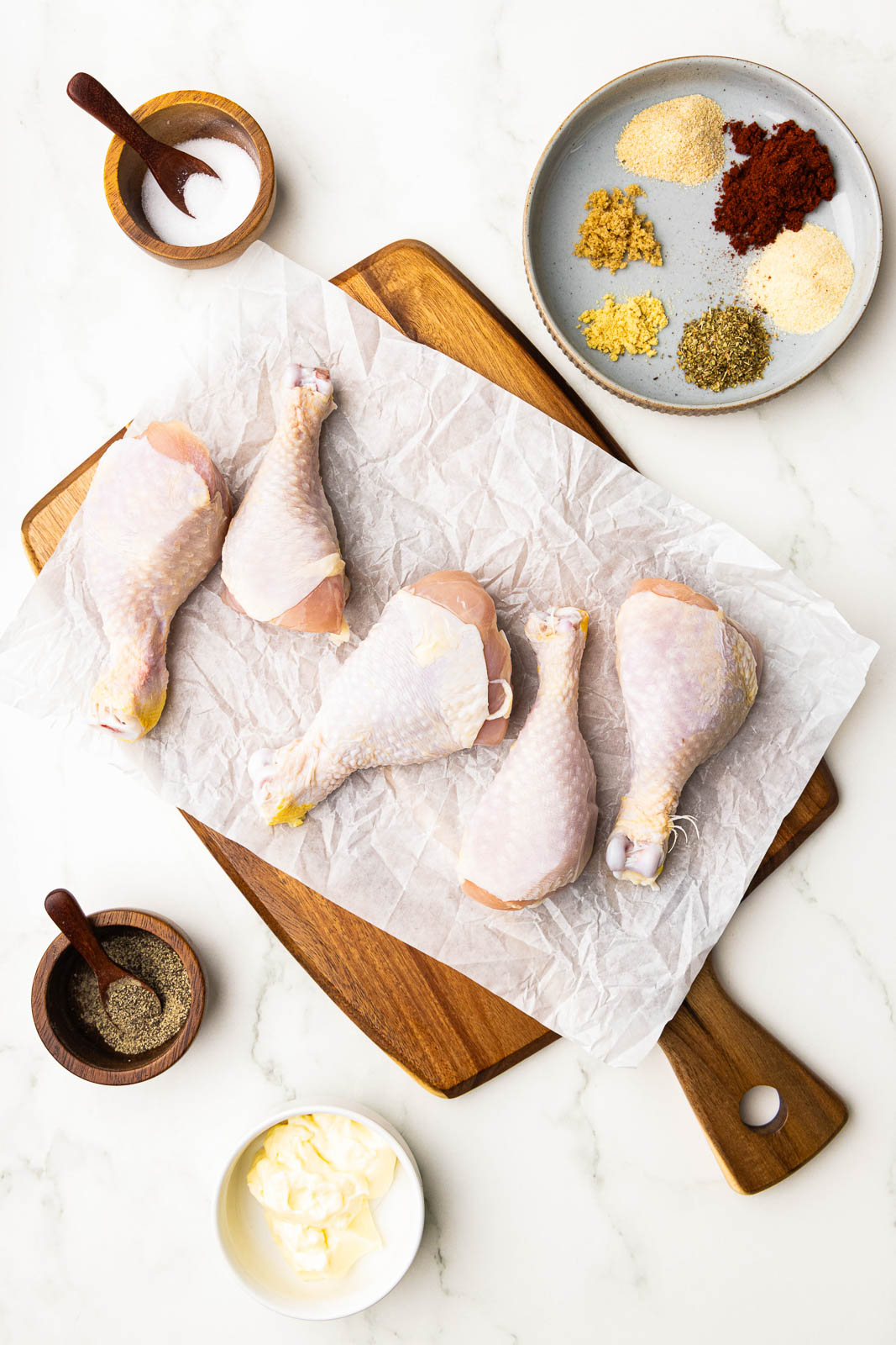 parchment lined wooden cutting board containing 5 chicken drumsticks surrounded by containers of salt, pepper, mayonnaise, and larger bowl of assorted seasonings