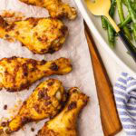 closeup view of parchment lined wooden cutting board containing five baked chicken drumsticks with platter of green beans and gold fork on side
