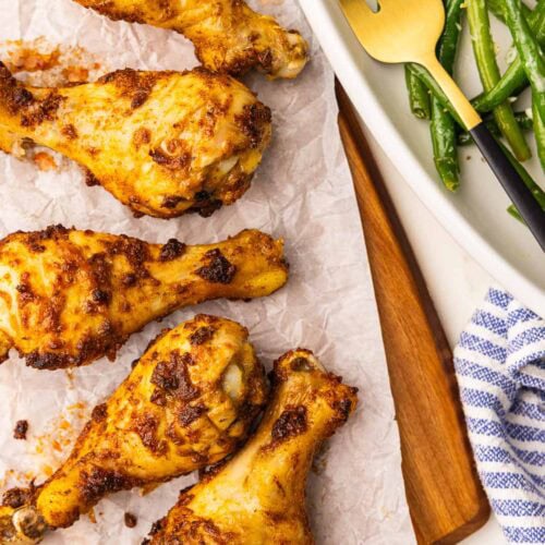 closeup view of parchment lined wooden cutting board containing five baked chicken drumsticks with platter of green beans and gold fork on side