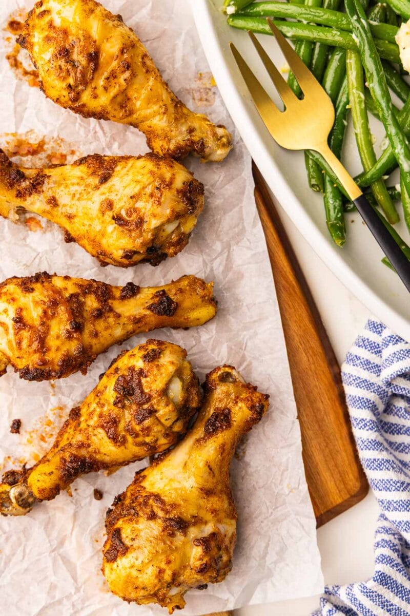 closeup view of parchment lined wooden cutting board containing five baked chicken drumsticks with platter of green beans and gold fork on side