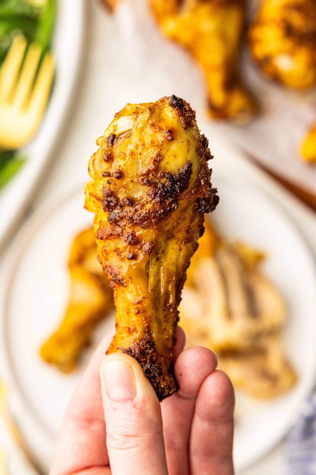 closeup view of hand holding baked chicken drumstick with blurred plates of chicken drumsticks and green beans in background