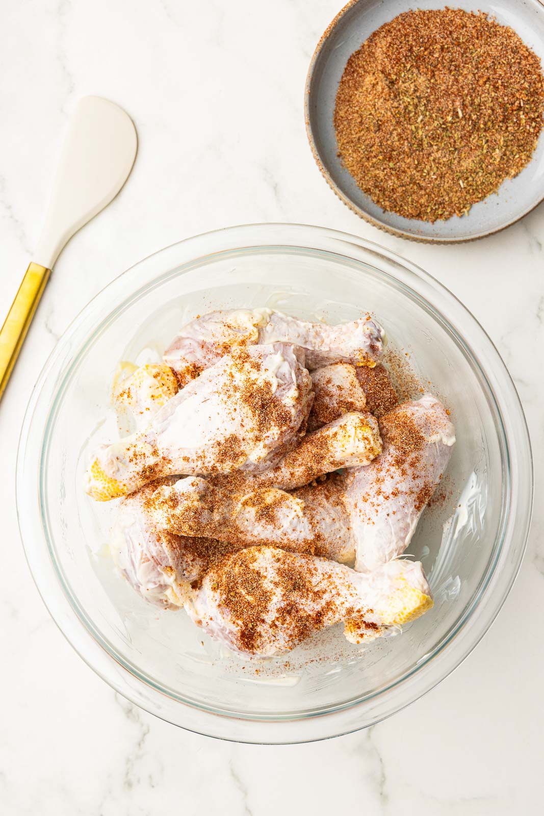 large glass bowl of mayonnaise coated chicken drumsticks sprinkled with seasonings, with bowl of seasonings and spatula on side