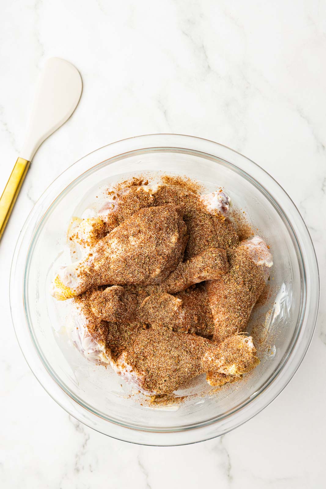 large glass bowl of chicken drumsticks coated with mayo and seasonings with spatula on side