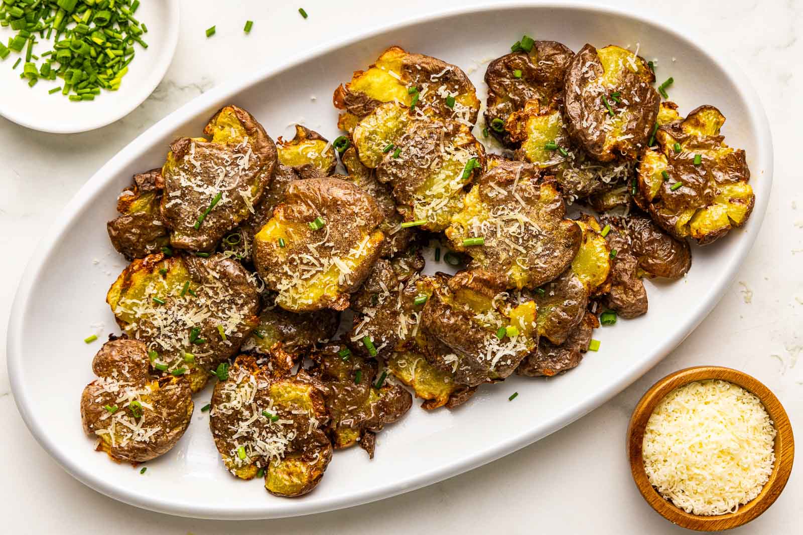 overhead side view of large white platter containing smashed potatoes sprinkled with Parmesan cheese and chives, with containers of Parmesan cheese and chives on the side