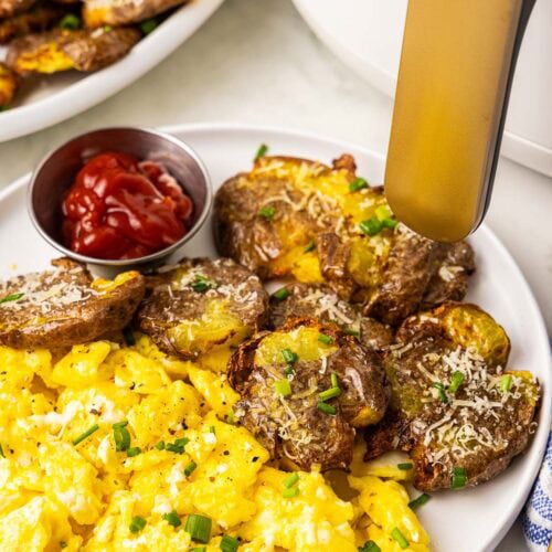 white dinner plate with smashed potatoes, scrambled eggs and ketchup with platter of potatoes and air fryer in background