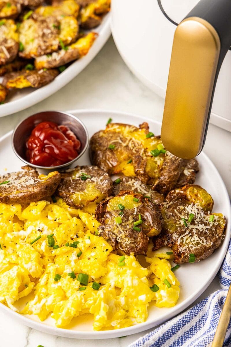white dinner plate with smashed potatoes, scrambled eggs and ketchup with platter of potatoes and air fryer in background