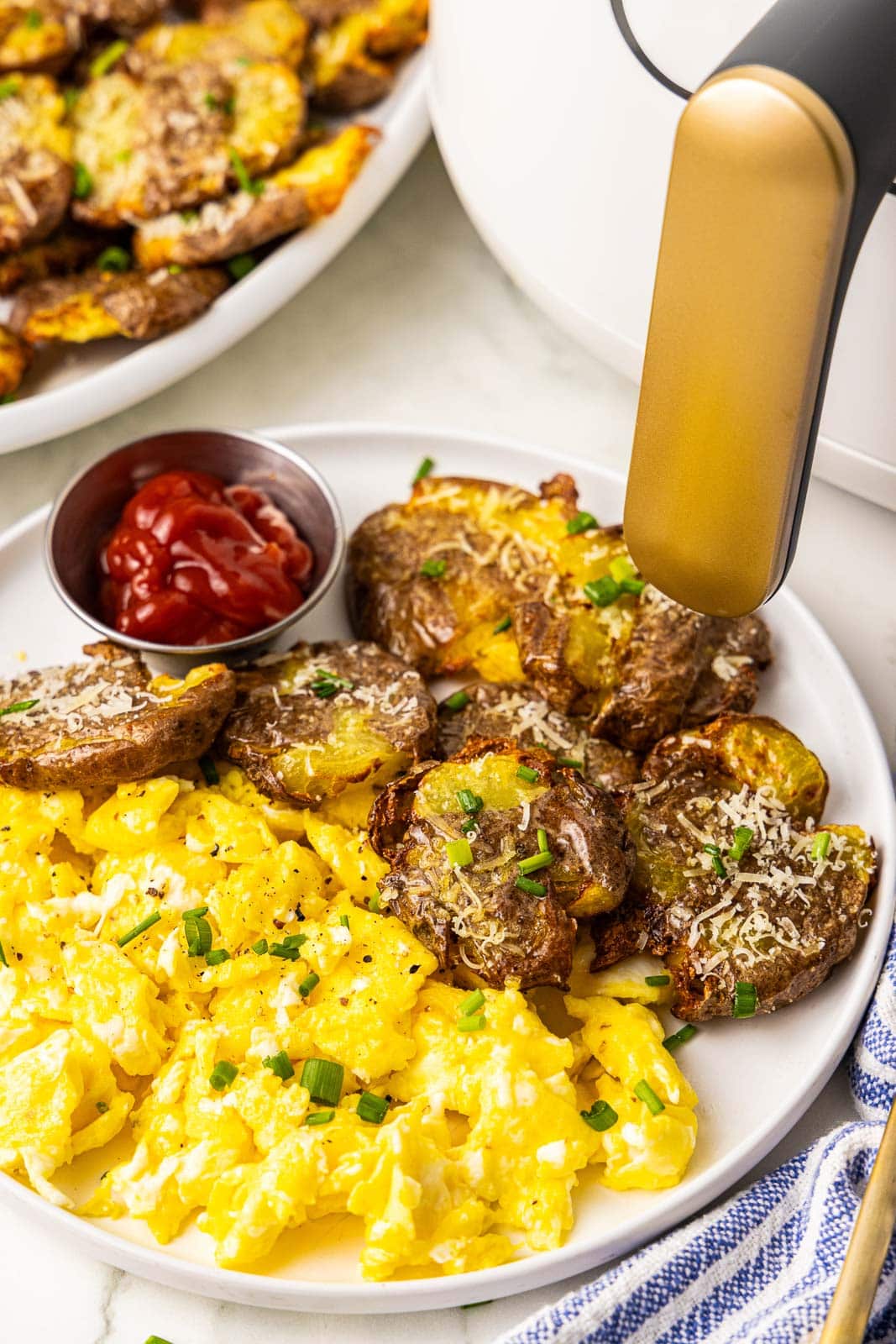 white dinner plate with smashed potatoes, scrambled eggs and ketchup with platter of potatoes and air fryer in background