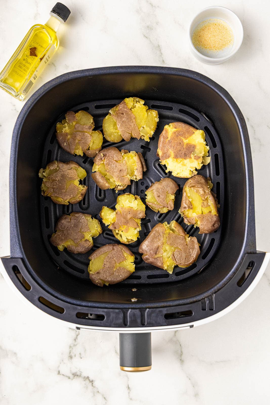 overhead view of black air fryer containing smashed potatoes with containers of olive oil and seasonings on side