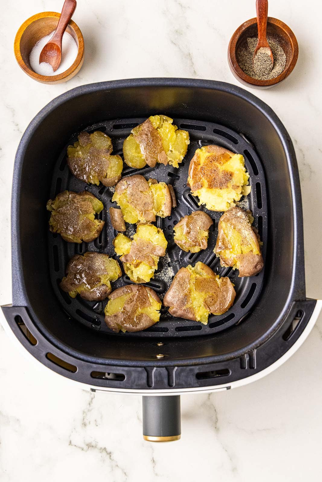 overhead view of black air fryer containing smashed potatoes that have been oiled with salt and pepper containers on side