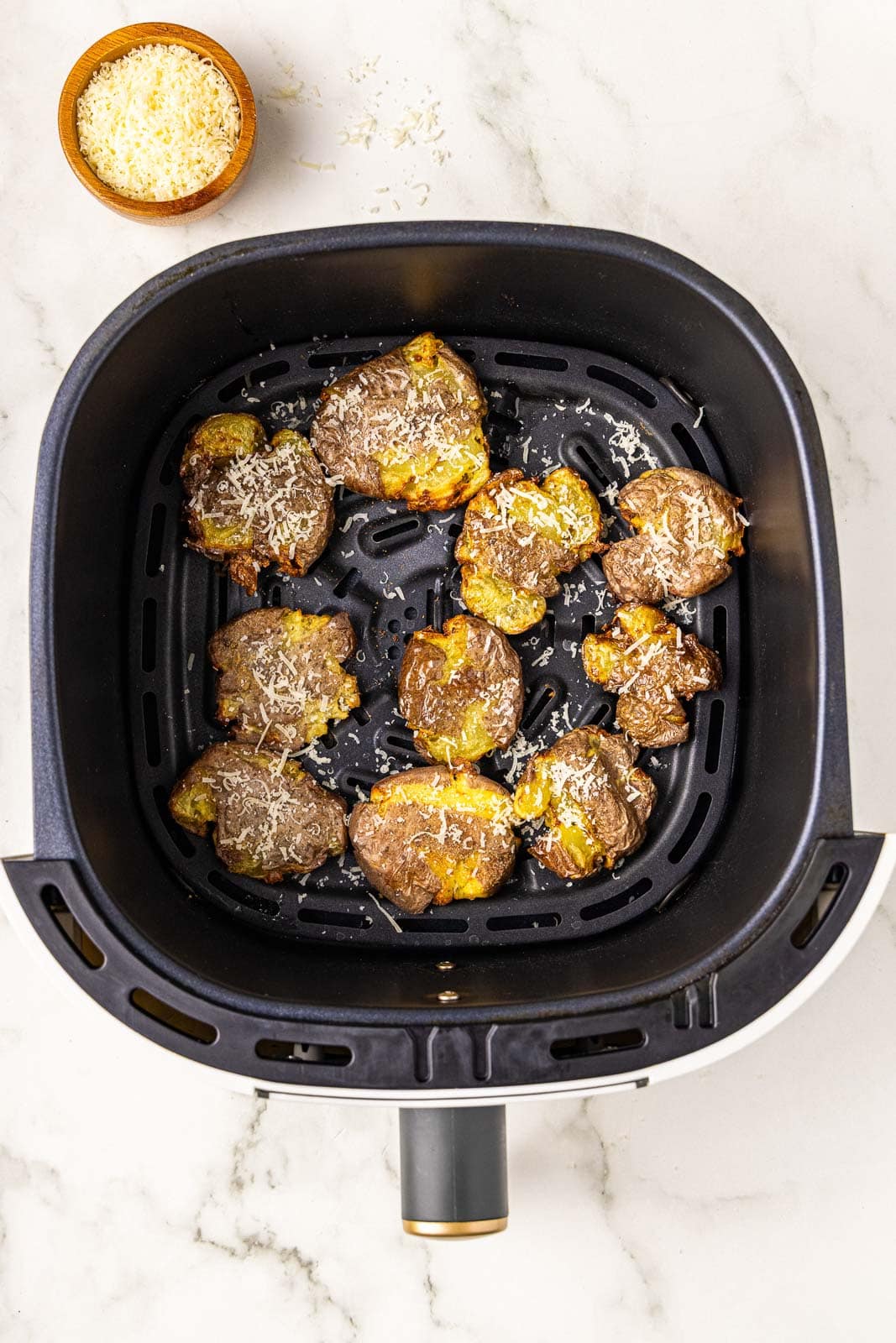 overhead view of air fryer with smashed potatoes with Parmesan cheese on top and container of Parmesan cheese on the side