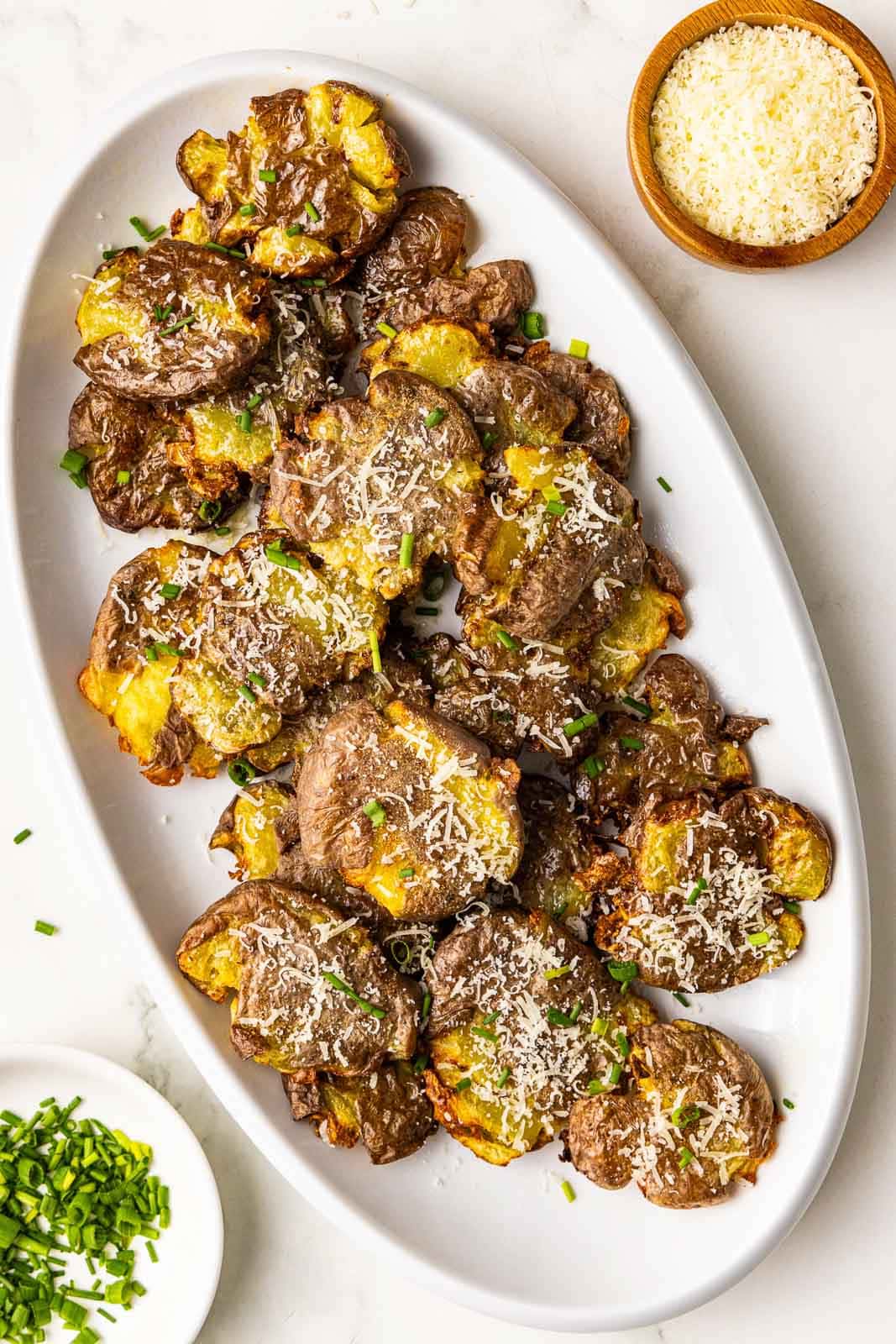 large white platter containing smashed potatoes sprinkled with Parmesan cheese and chives, with containers of Parmesan cheese and chives on the side