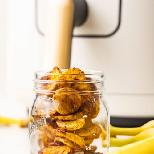 glass jar containing air fried banana chips with bananas and air fryer in background