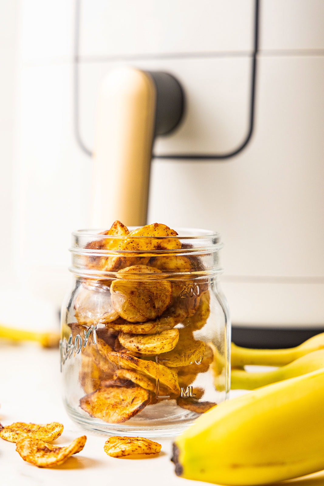 glass jar containing air fried banana chips with bananas and air fryer in background