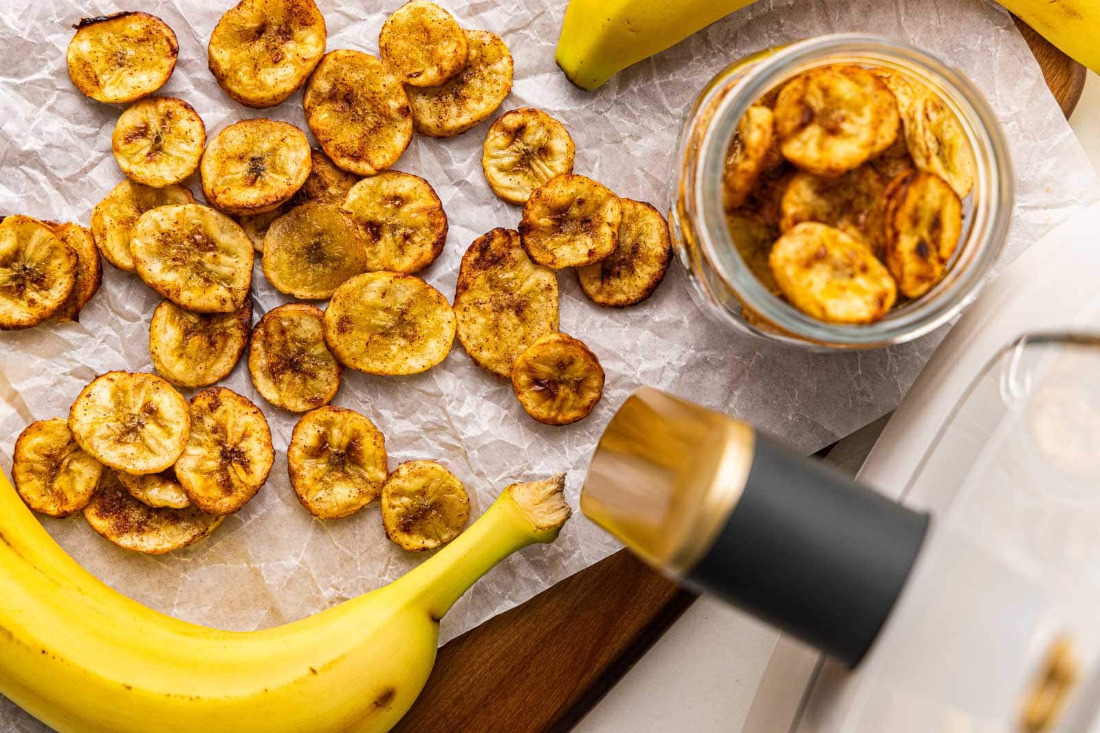 parchment lined wooden cutting board containing air fried banana chips, glass jar of air fried banana chips, with bananas and air fryer in the background