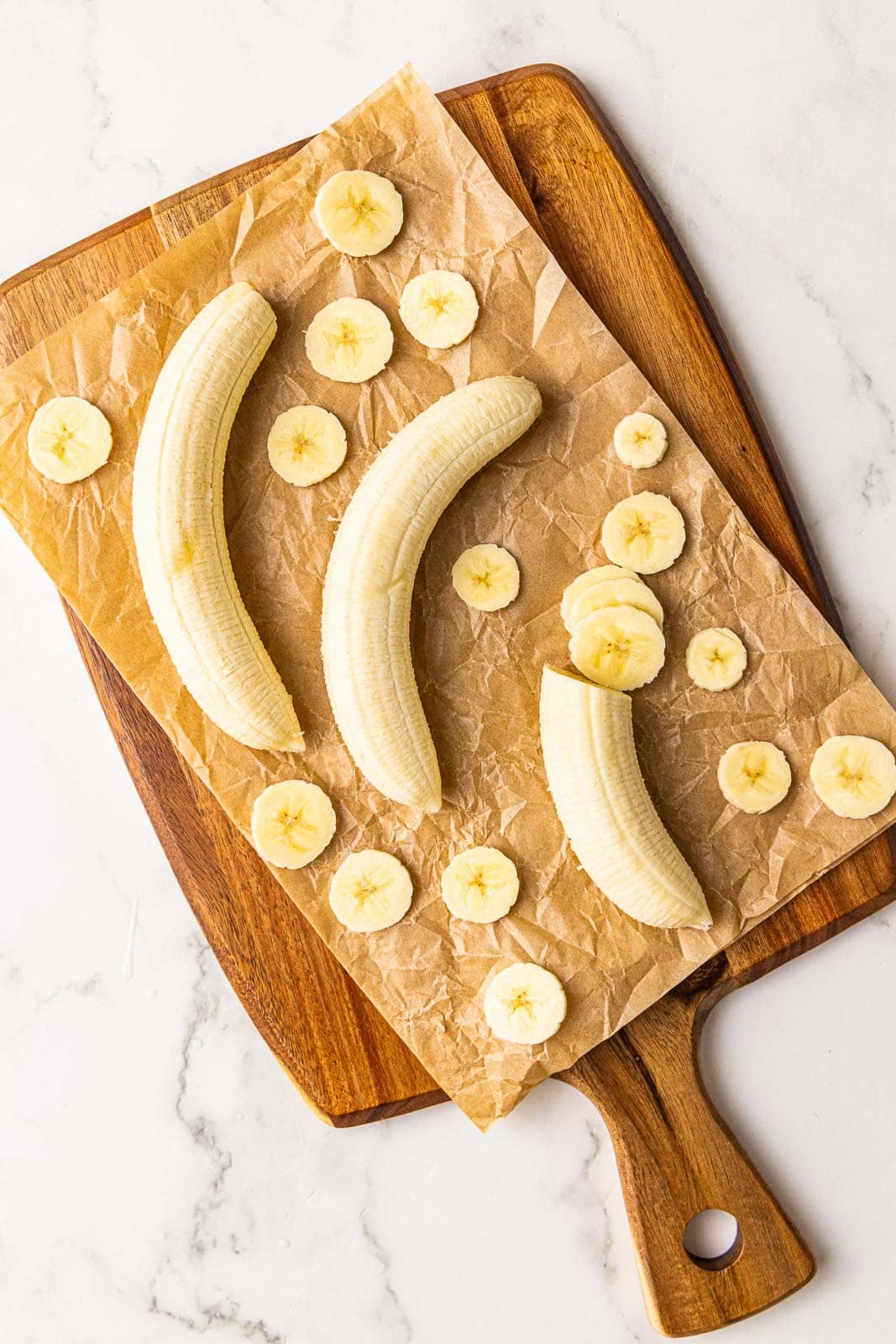 parchment lined wooden cutting board containing 3 bananas with some having been sliced thinly