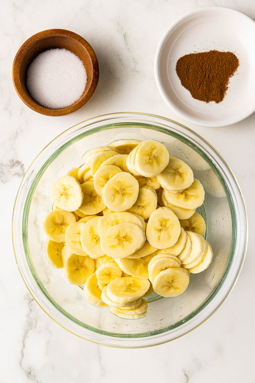 bowl of sliced bananas that have been tossed with lemon juice with bowls of sugar and cinnamon on side
