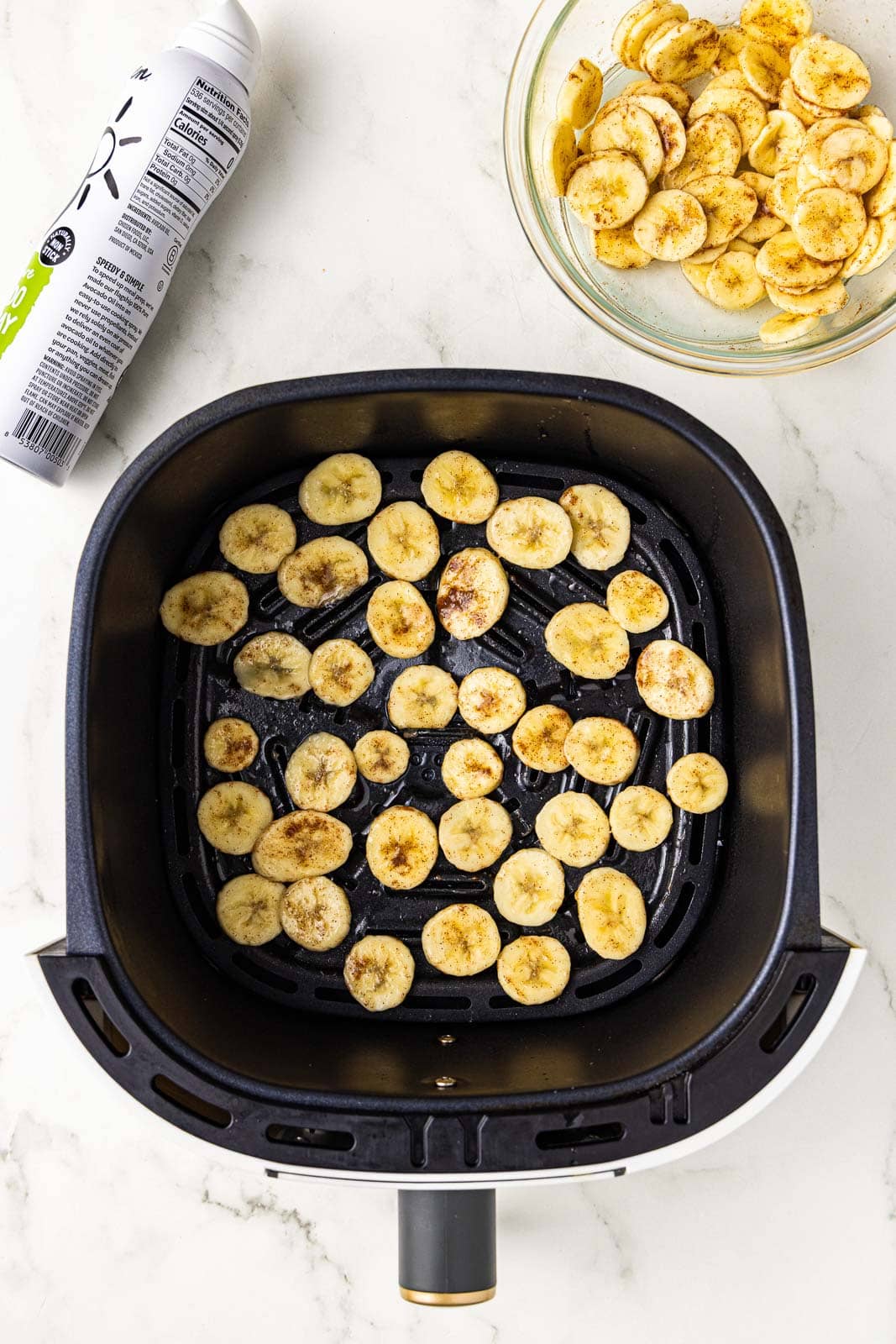 overhead view of black air fryer with sliced bananas with bowl of bananas and avocado spray on side