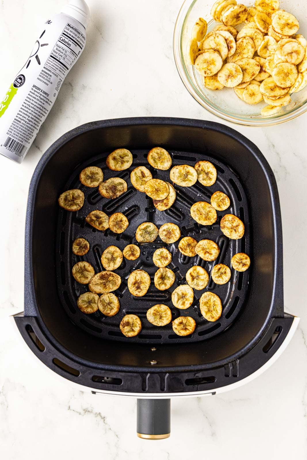 overhead view of black air fryer with air fried sliced bananas with bowl of bananas and avocado spray on side