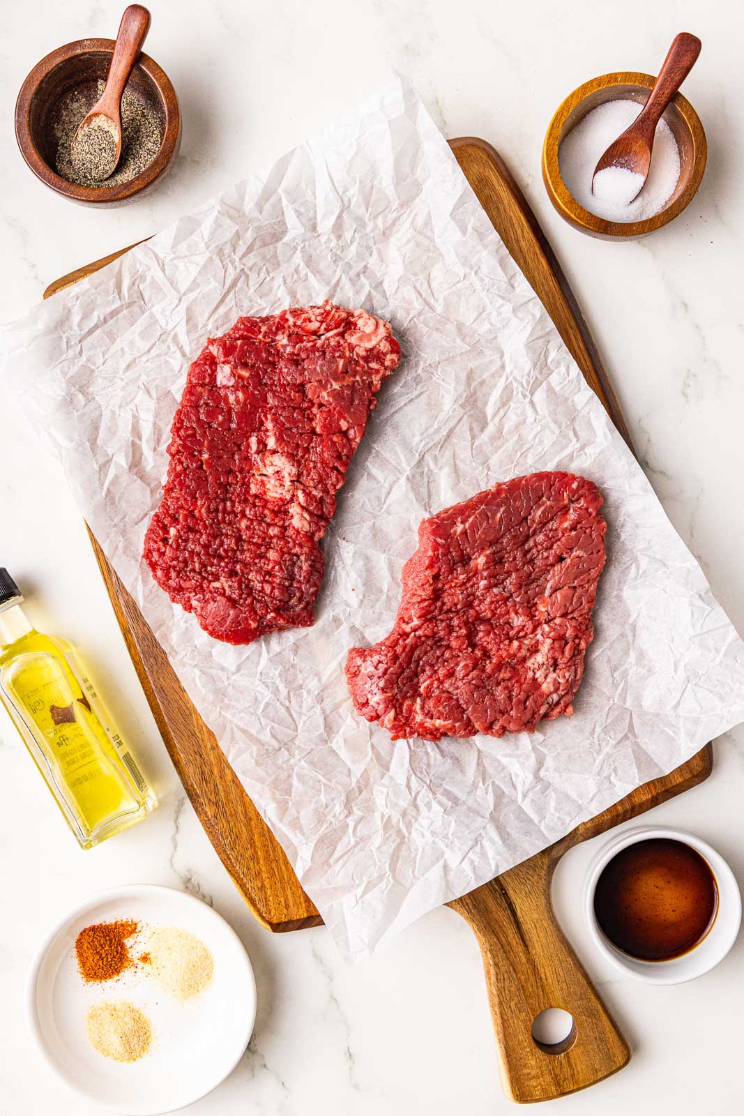 ingredients to include parchment lined wooden cutting board with 2 cube steaks surrounded by containers of olive oil, Worcestershire sauce, salt, pepper, garlic powder, onion powder and smoked papria