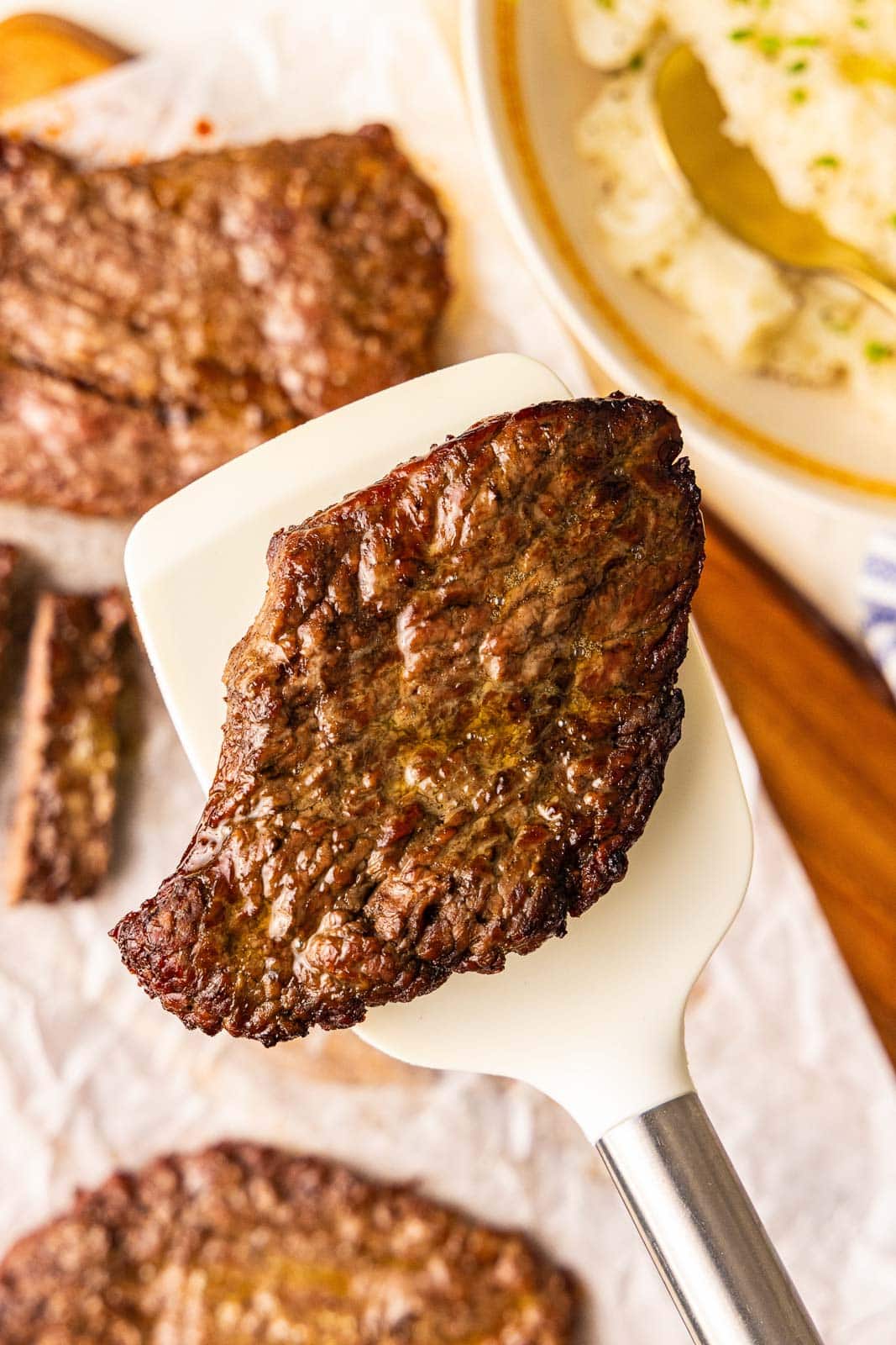 closeup view of air fried cube steak with parchment lined wooden cutting board with cube steak and bowl of mashed potatoes in background