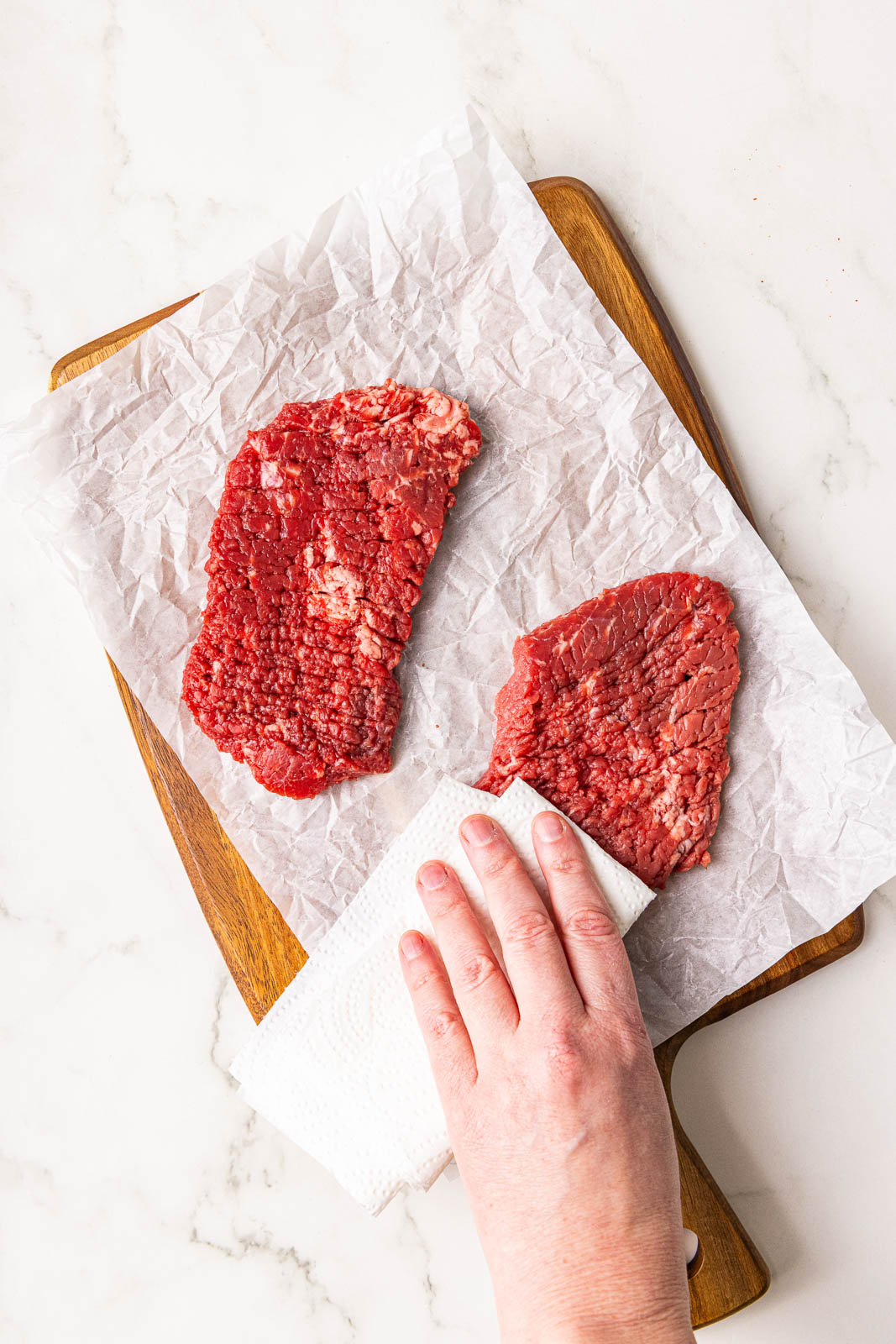 parchment lined wooden cutting board with hand patting 2 cube steaks dry with paper towel