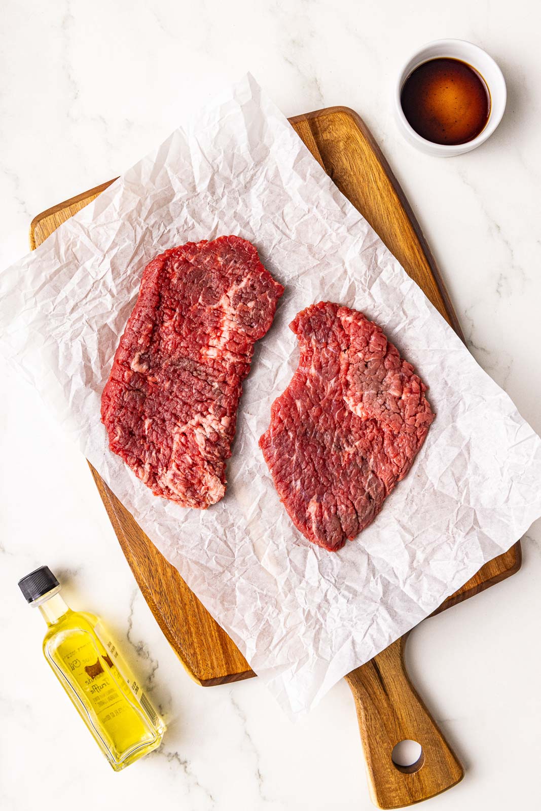 parchment lined wooden cutting board with 2 cube steaks surrounded by olive oil and Worcestershire sauce