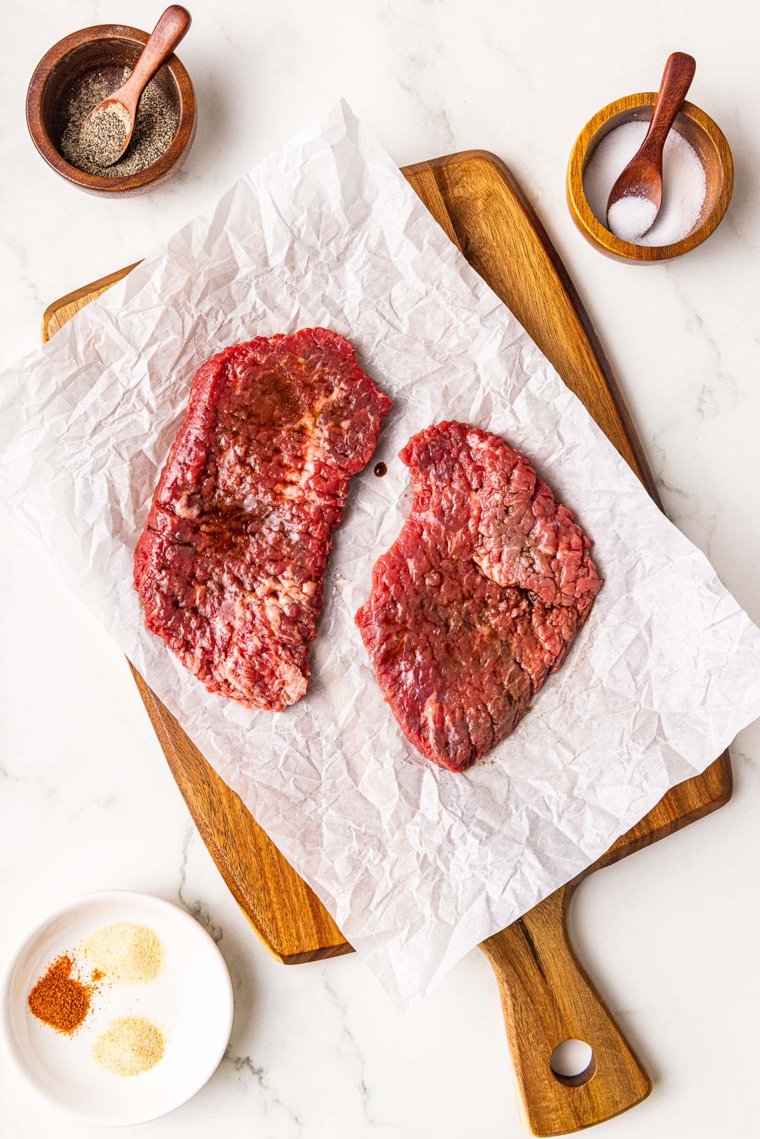 parchment lined wooden cutting board with 2 cube steaks surrounded by containers of salt, pepper and assorted seasonings