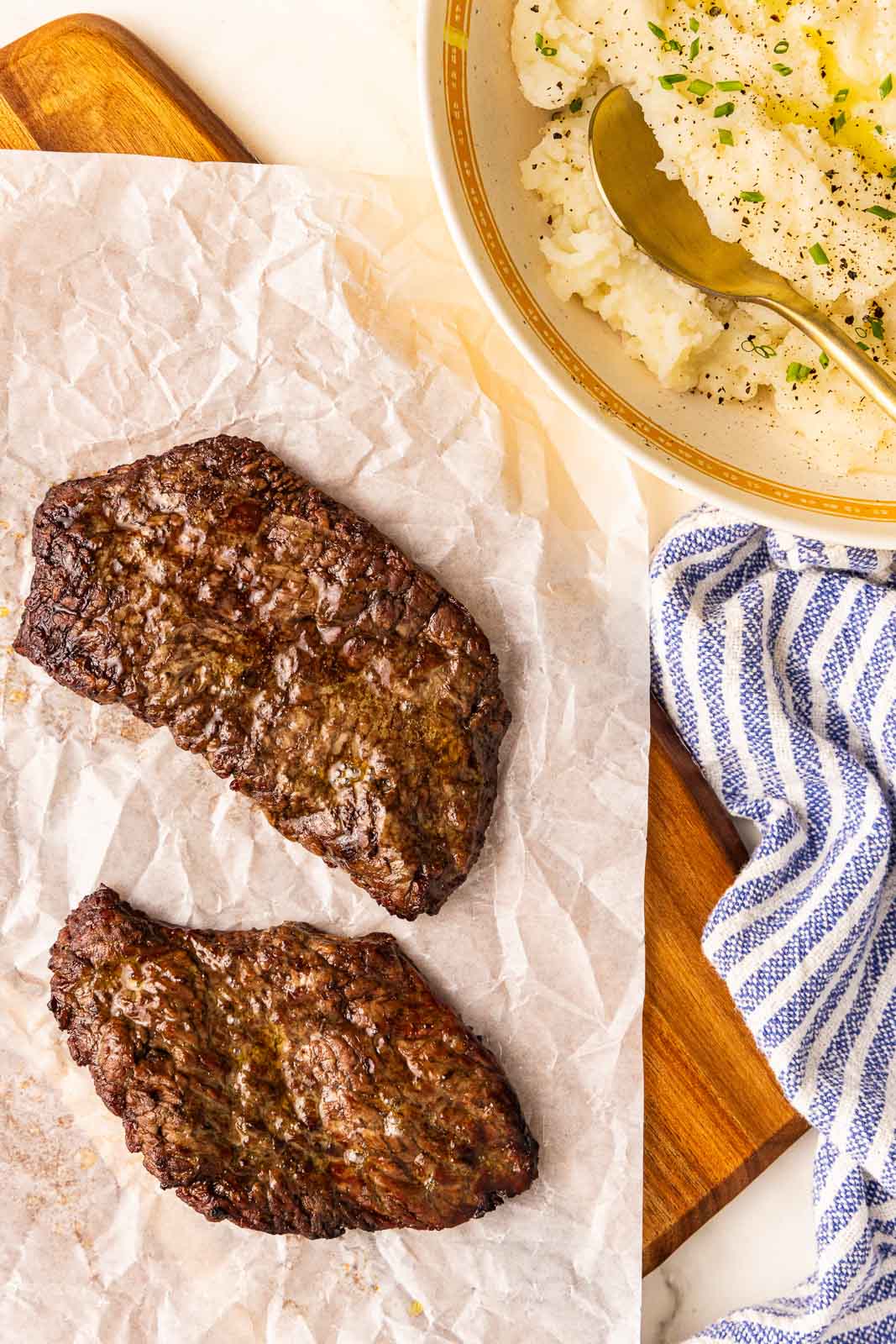 parchment lined wooden cutting board with 2 air fried cube steaks with bowl of mashed potatoes on side