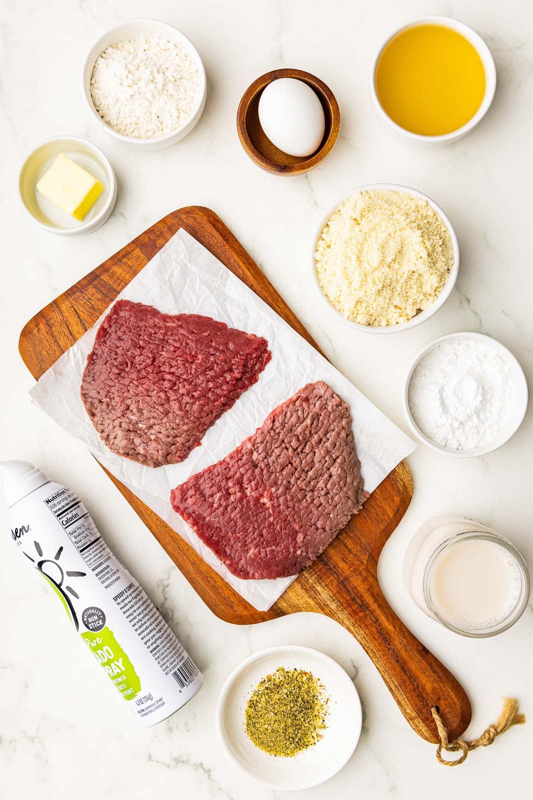 ingredients to include parchment lined wooden cutting board with 2 pieces of cube steak surrounded by containers of egg, almond flour, tapioca flour, her and garlic seasoning, avocado spray, butter, gluten free flour, chicken broth and almond milk