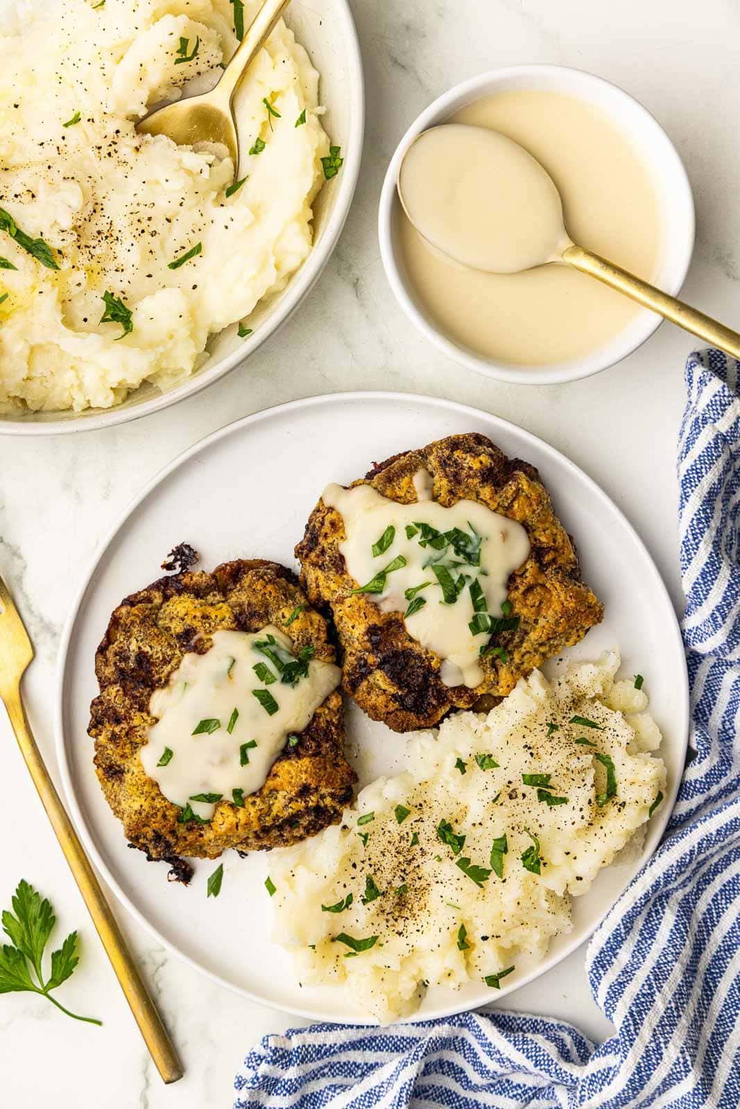 white dinner plate with 2 pieces of cube steak, mashed potatoes with parsley, bowl of mashed potatoes, gravy with spoon and fork on the side