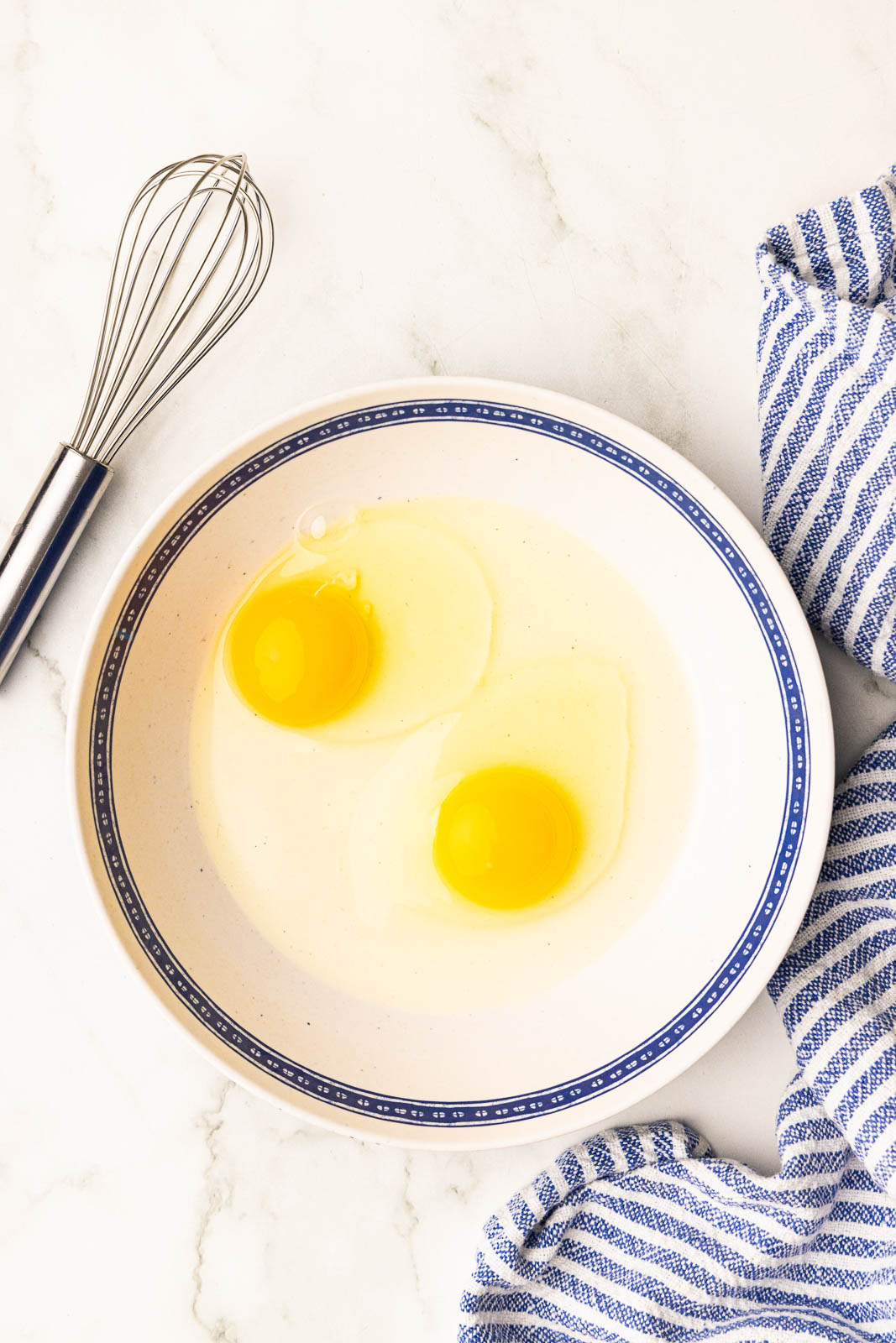 overhead view of large white bowl with 2 eggs with a wire whisk on the side