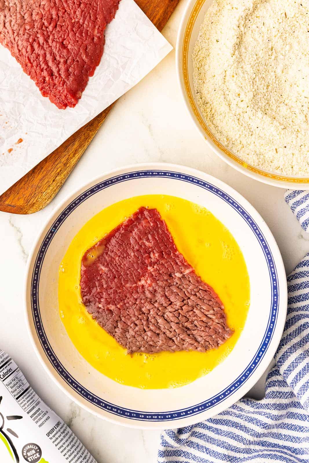 large bowl with beaten egg and one cube steak being dipped in mixture, with another container of flour mixture and wooden cutting board with cube steak on side