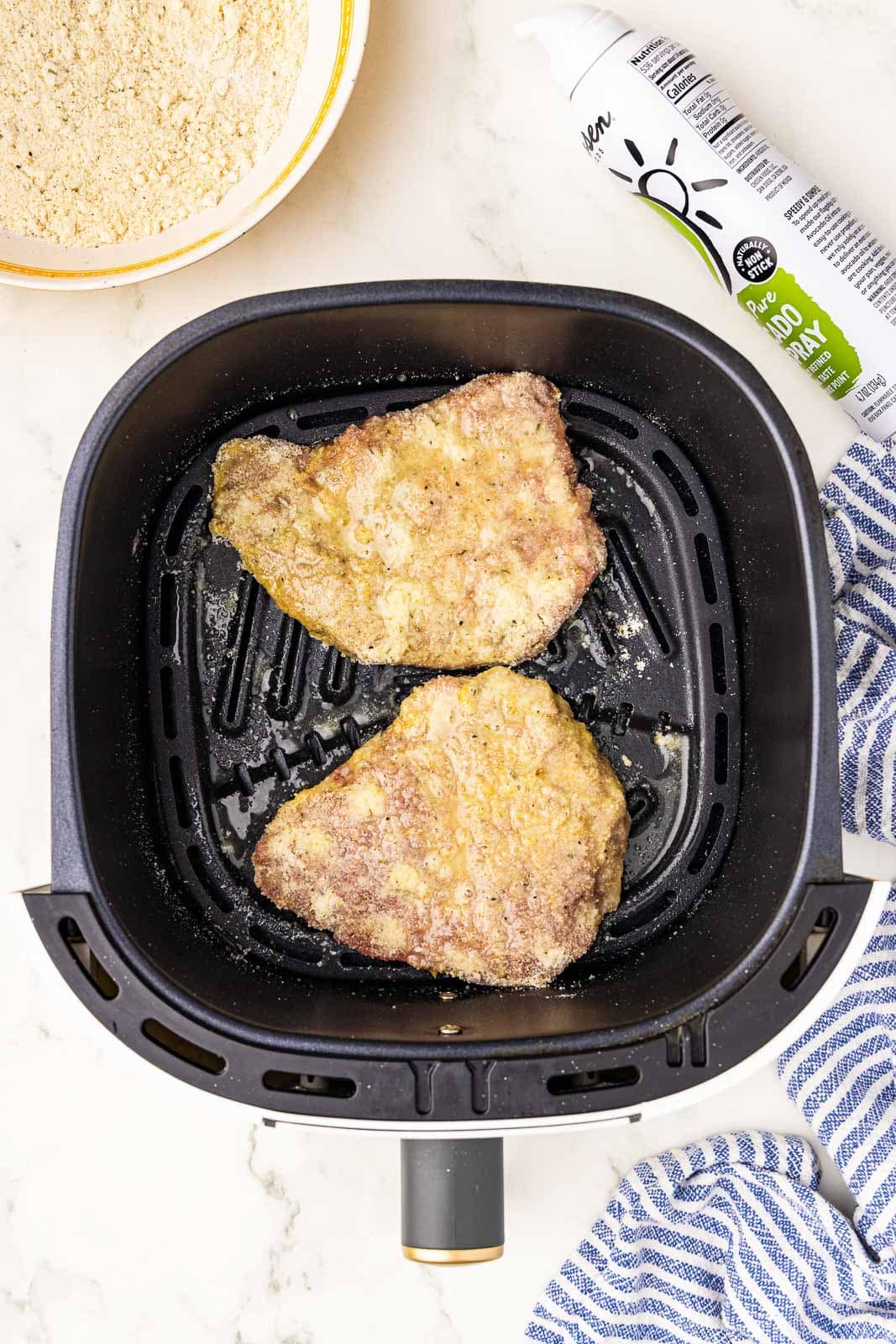 overhead view of black air fryer with 2 cube steak, avocado spray and bowl with flour mix on side