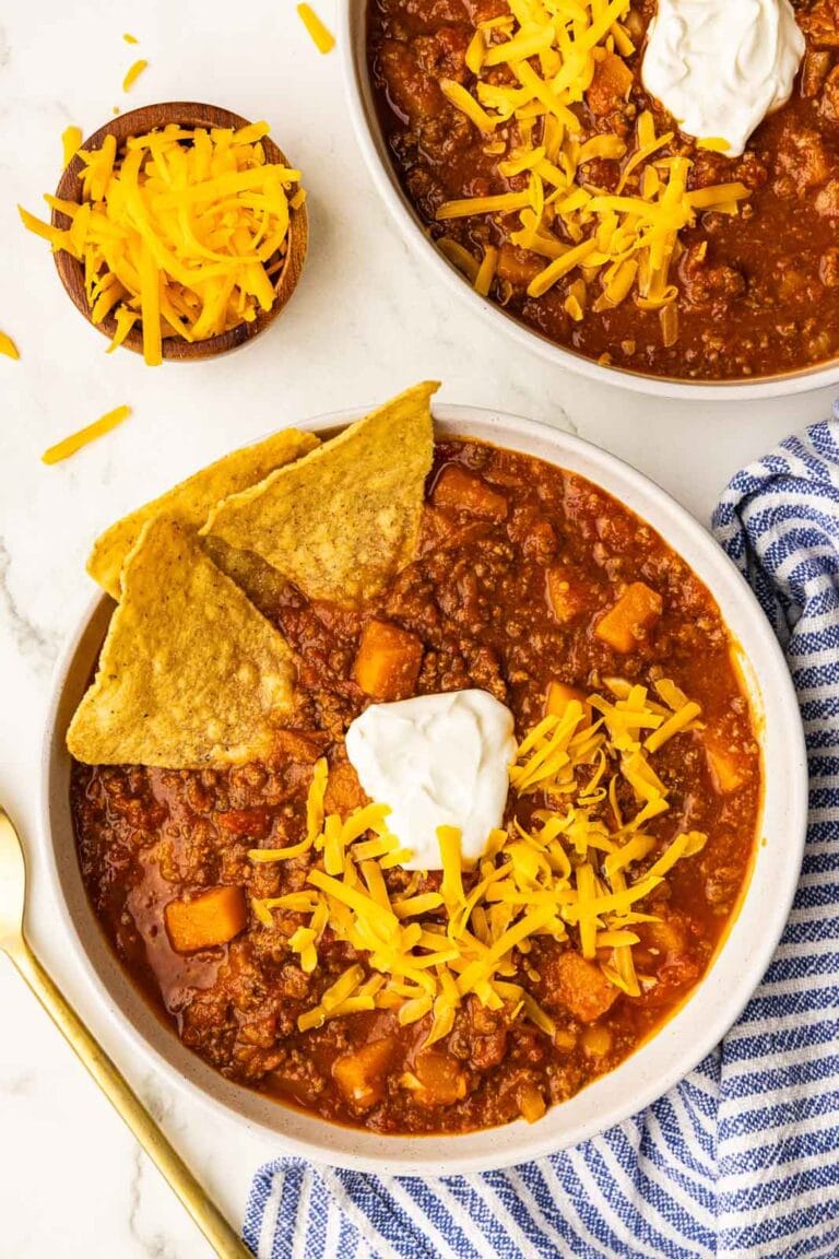 two bowls of chili with dollop of sour cream, cheese and tortilla chips, with bowl of cheese on side