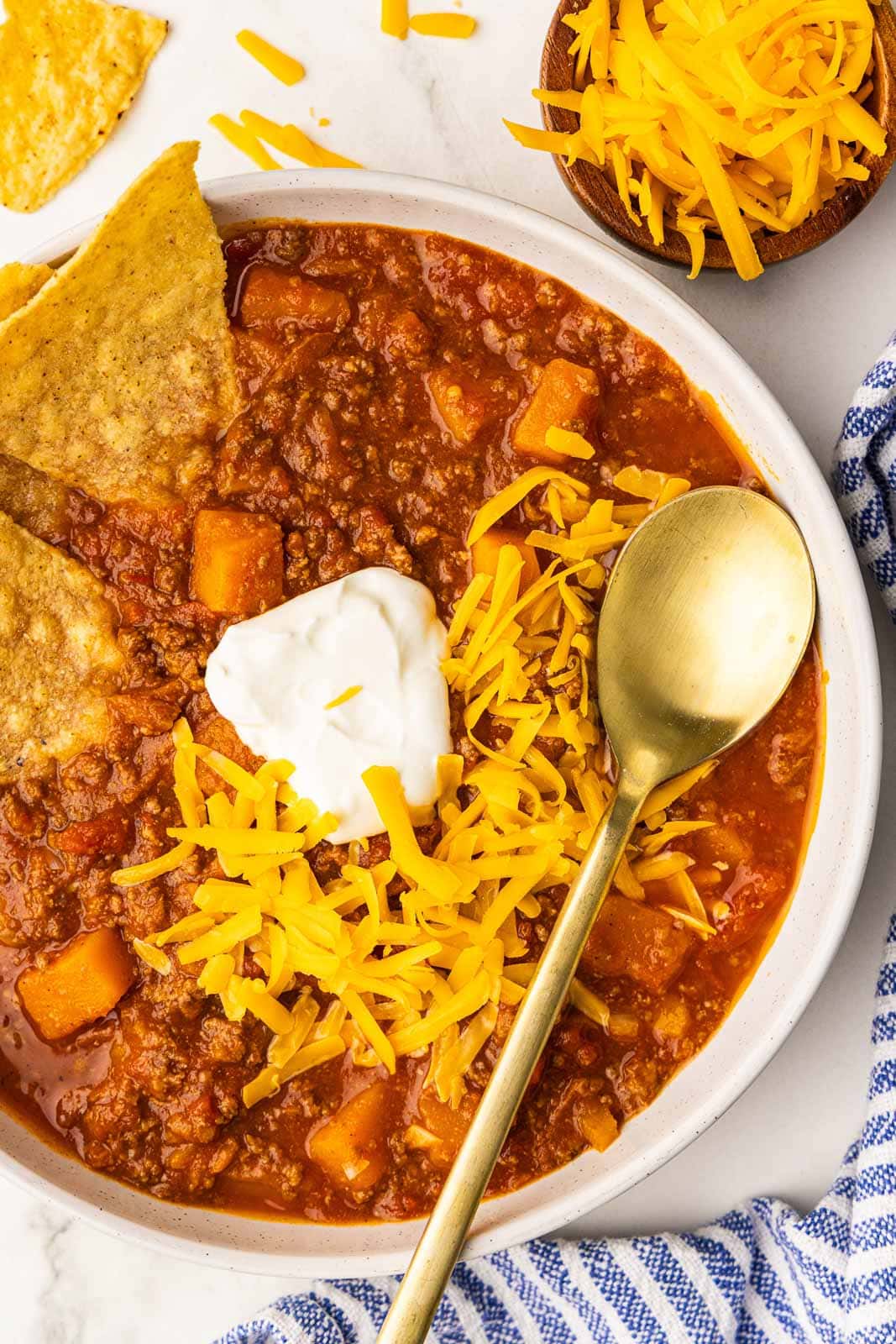closeup of white bowl of chili with sour cream, cheese and tortilla chips, with bowl of cheese on side