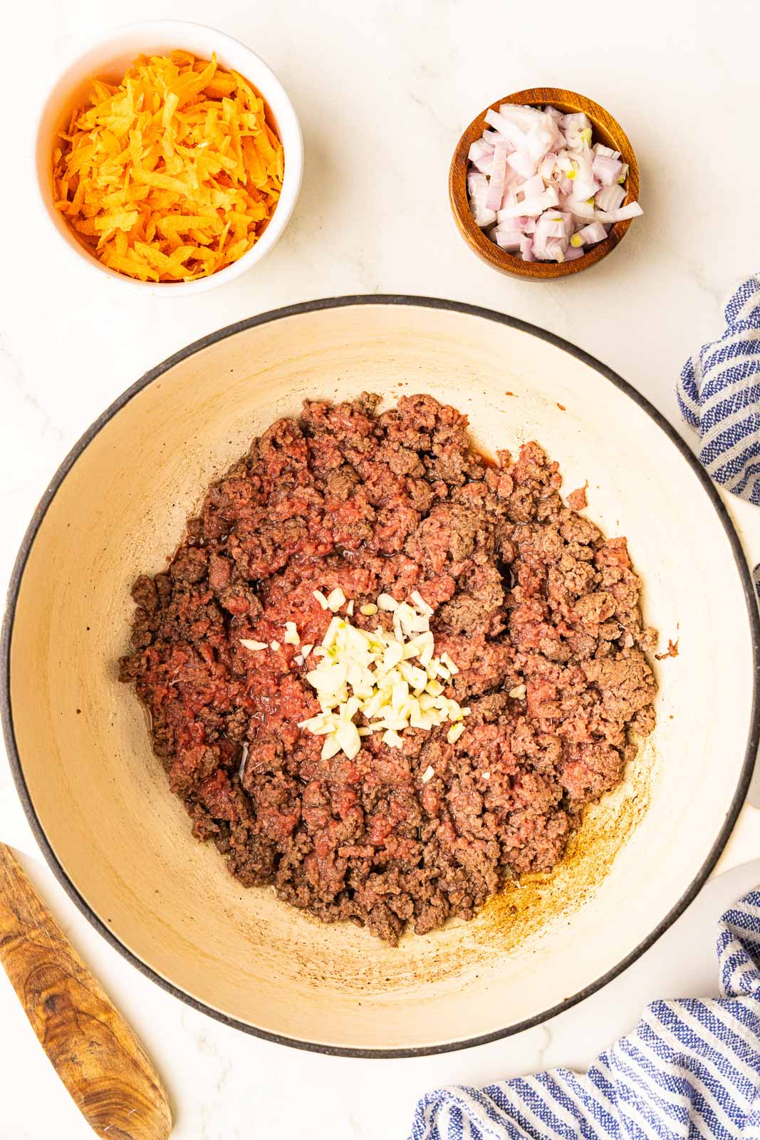 overhead view of Dutch oven with partially browned ground bison with garlic on top and containers of carrot and shallot on the side