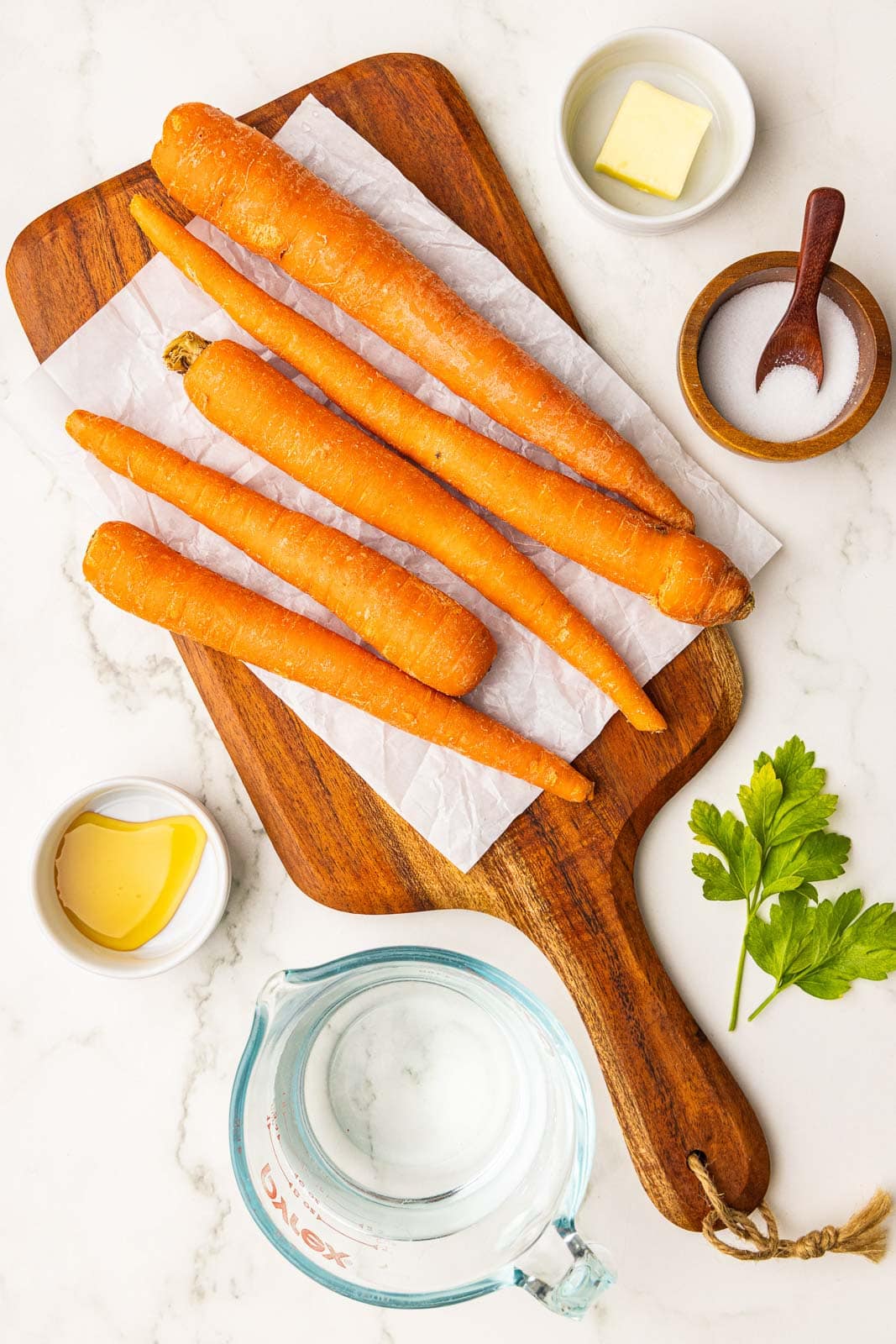 ingredients to include parchment lined wooden cutting board with 5 carrots surrounded by containers of butter, salt, water, and honey