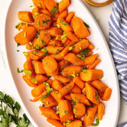 overhead view of white platter of cooked carrots with parsley on top, with bowl of salt and parsley leaves on the side