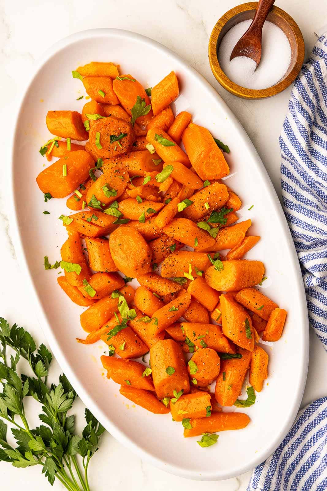 overhead view of white platter of cooked carrots with parsley on top, with bowl of salt and parsley leaves on the side