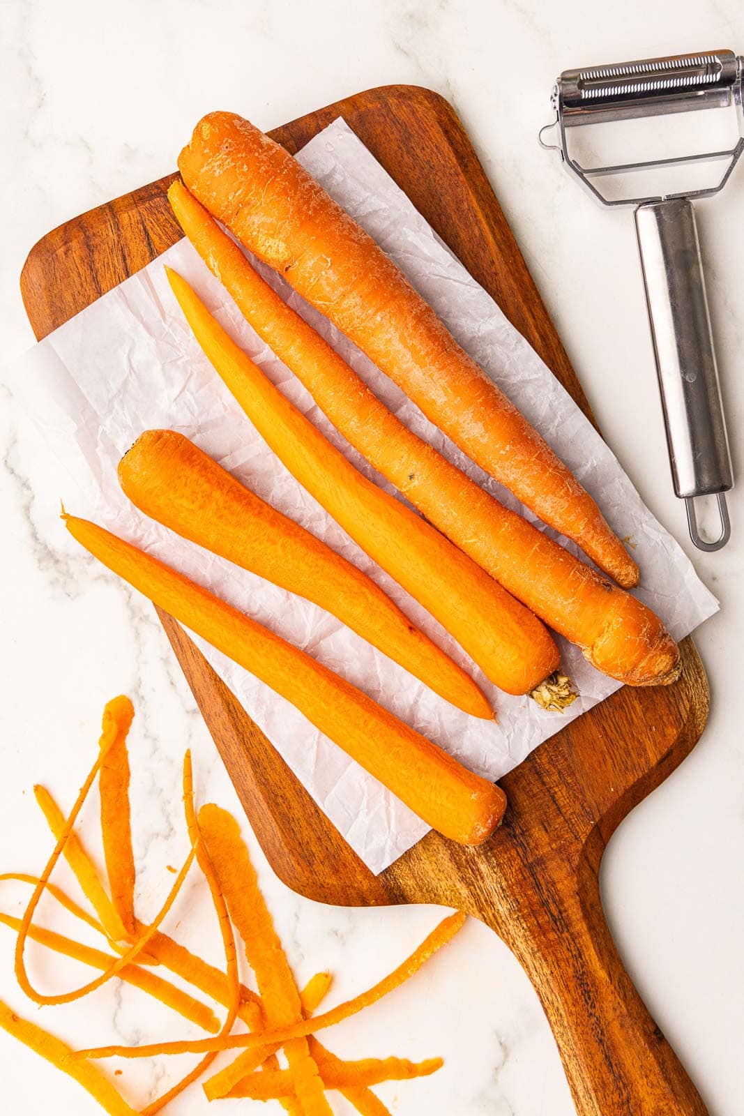 parchment lined wooden cutting board with carrots 5 carrots, 3 of which are peeled, with peeler on side