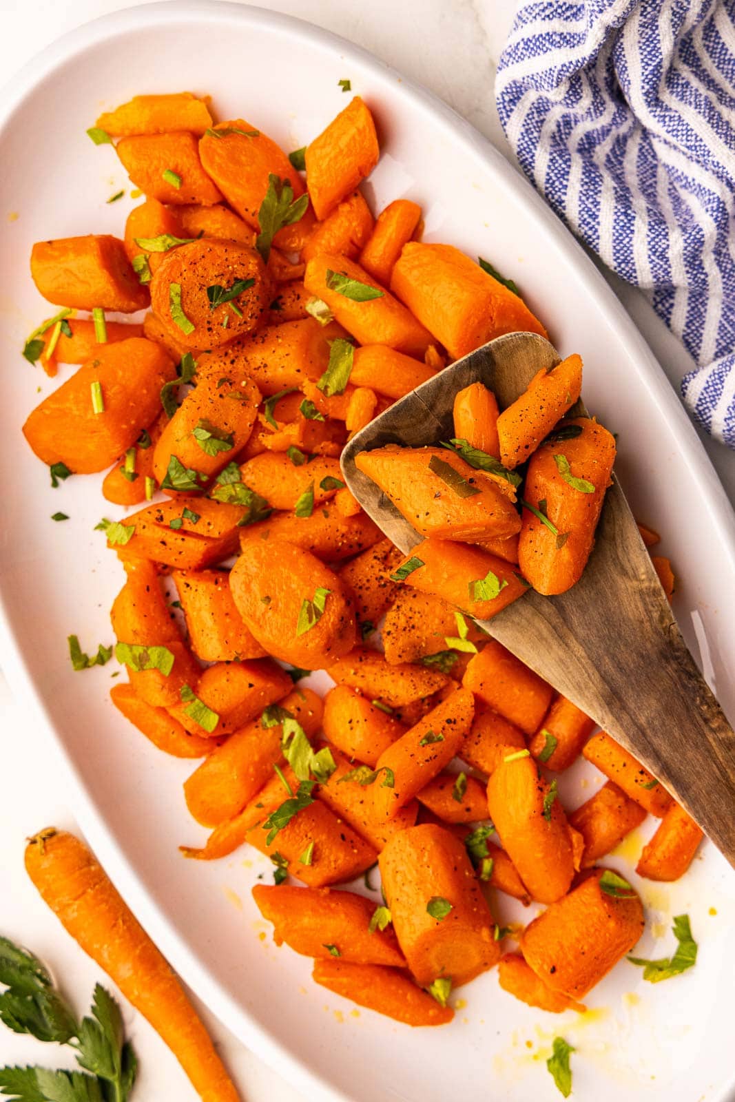 overhead view of cooked carrots sprinkled with parsley on white platter with wooden spoon, with a raw carrots and parsley on side