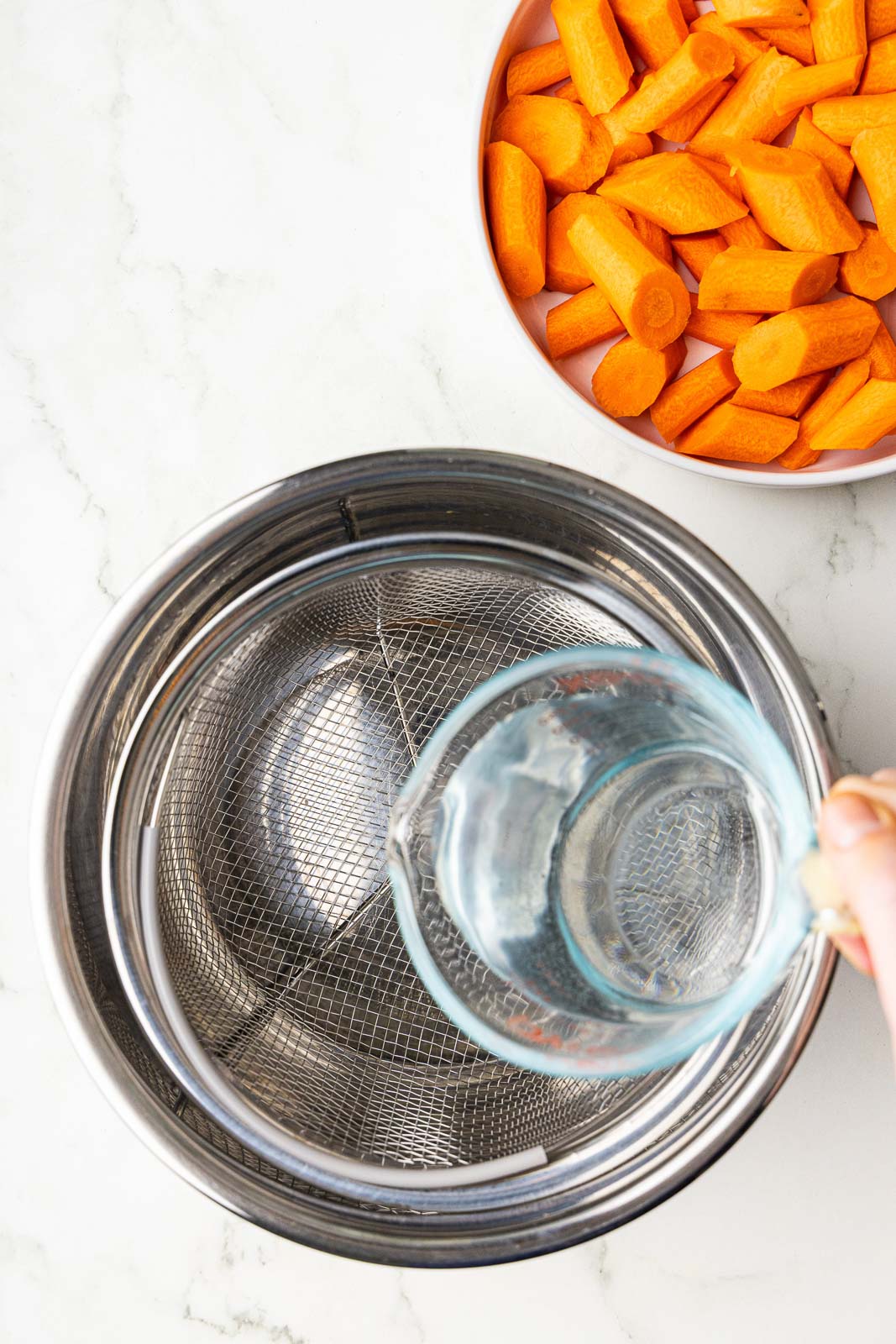 instant pot bowl and basket with hand pouring water into bowl with white bowl of wedged carrots on side