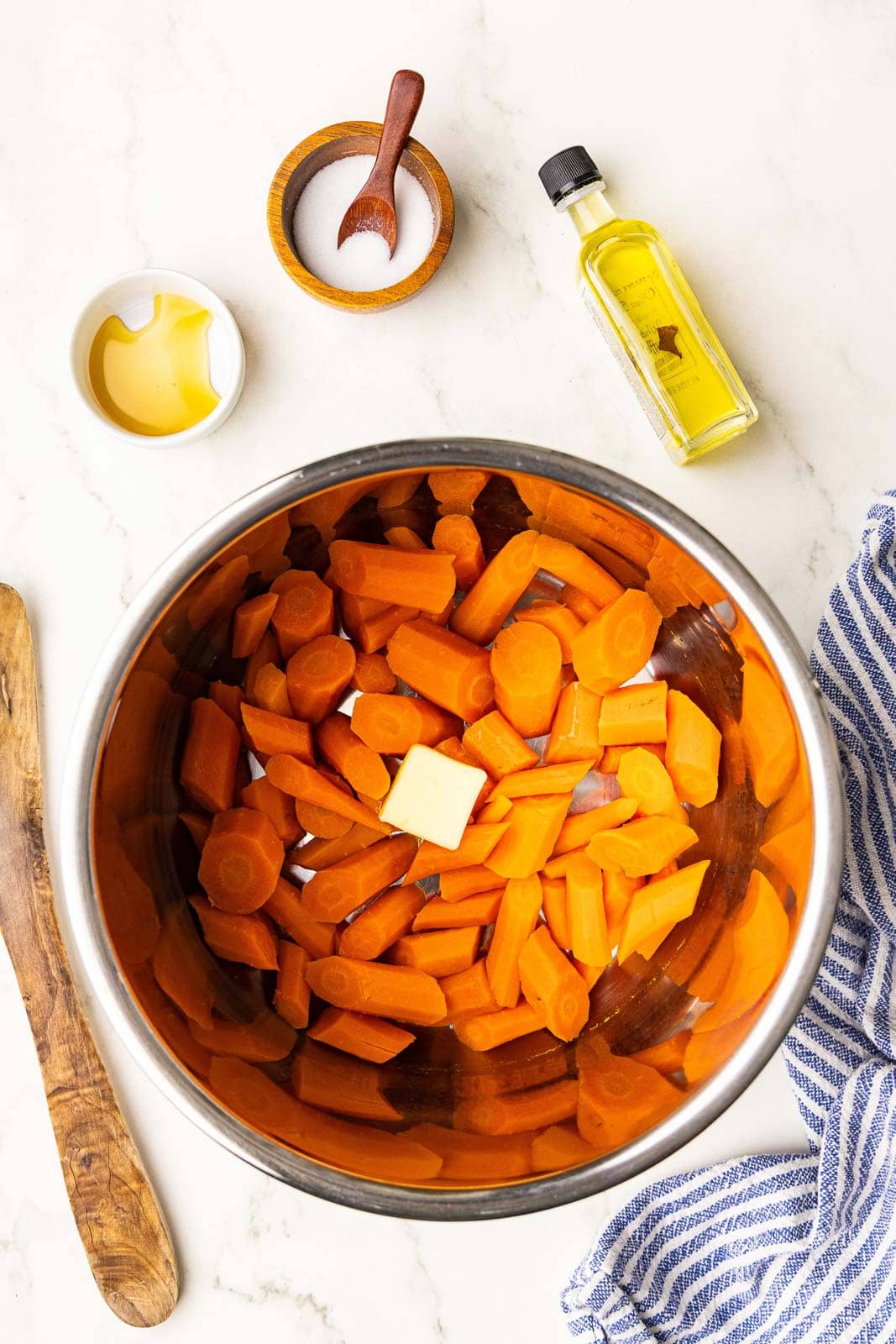 overhead view of cooked carrots in bowl with pat of butter on top surrounded by olive oil, salt, pepper and wooden spoon