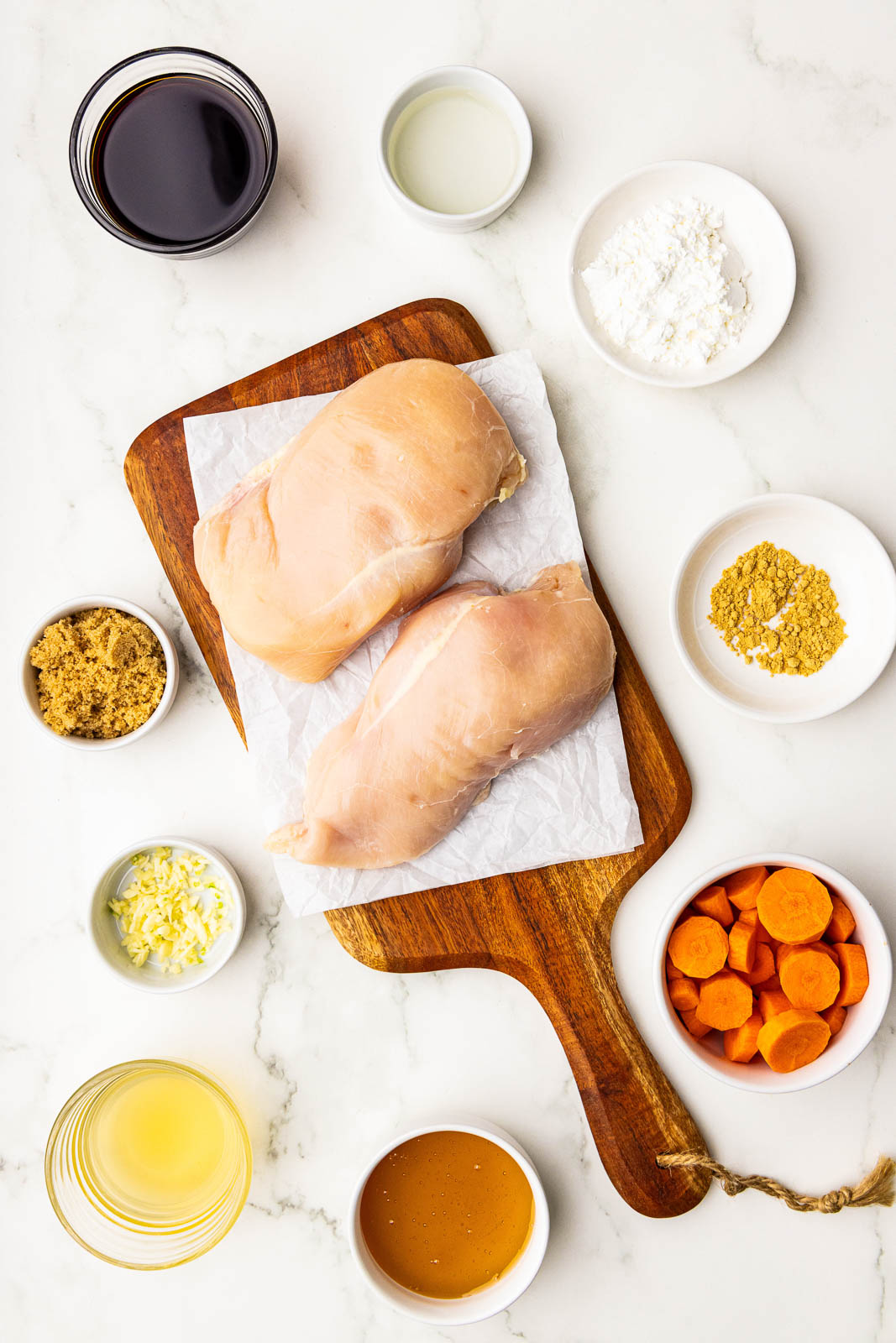 ingredients to include parchment lined wooden cutting board with 2 chicken breasts, surrounded by containers of soy sauce, water, corn starch, ground ginger, sliced carrots, honey, chicken broth, ground ginger and brown sugar
