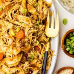 large white platter of chicken and carrots garnished with scallions and sesame seeds with fork, surrounded by bowls of white rice, scallions and sesame seeds