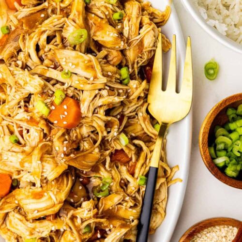 large white platter of chicken and carrots garnished with scallions and sesame seeds with fork, surrounded by bowls of white rice, scallions and sesame seeds