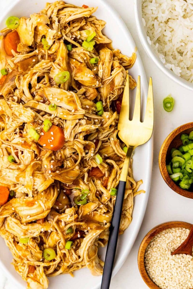 large white platter of chicken and carrots garnished with scallions and sesame seeds with fork, surrounded by bowls of white rice, scallions and sesame seeds
